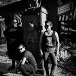 The band is in front of what looks like a grave marker with a gargoyle on top. Wyatt is standing to the left of the monument, Tommy is in the forefront squatting, and Mimi is to the right.